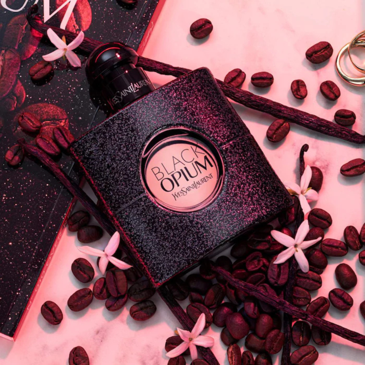 Yves Saint Laurent Black Opium perfume bottle surrounded by coffee beans and flowers on a pink background
